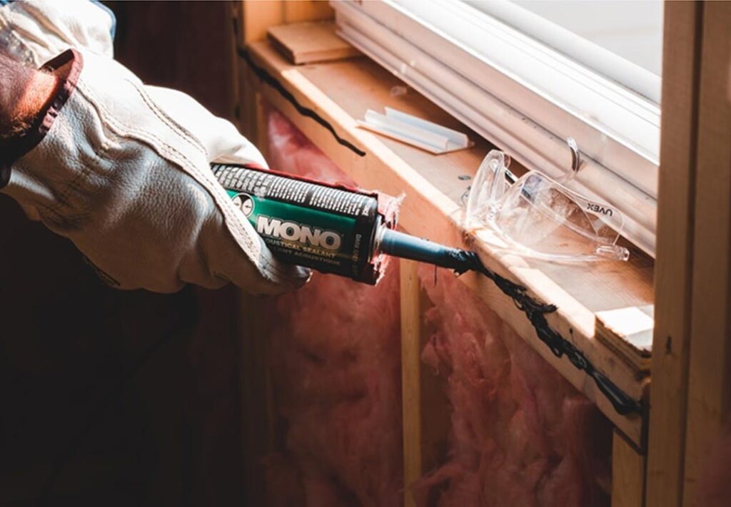 Close-up of a gloved hand applying sealant to repair window damage, with protective glasses and insulation materials nearby, highlighting the process of restoring energy efficiency and structural integrity.